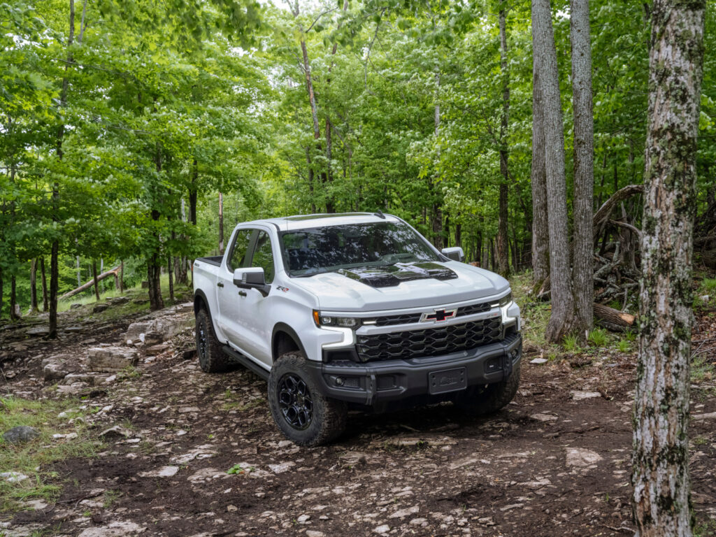 White pickup truck driving on a rugged forest trail, surrounded by lush green trees.