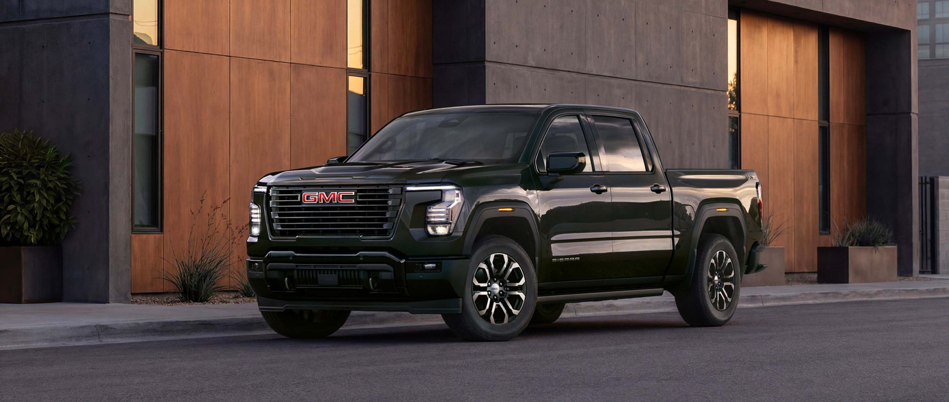 A green SUV is parked on a paved surface near a calm body of water. The car features a prominent grille and sleek design. In the background, hills are visible under a clear blue sky.