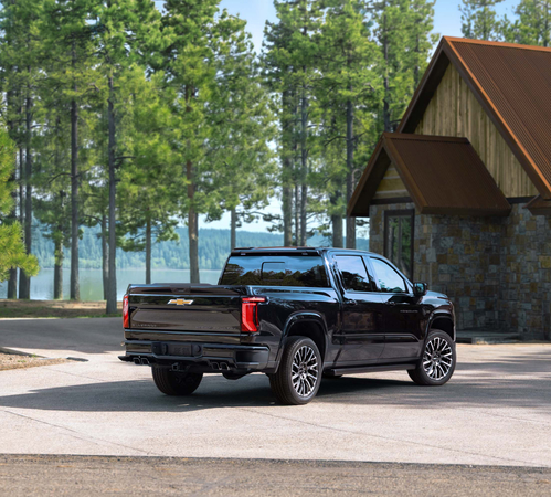 Black pickup truck parked near a rustic cabin with pine trees in the background.