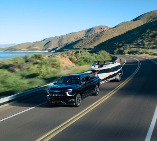 A black truck towing a boat on a scenic highway by mountains and a lake under a clear blue sky.