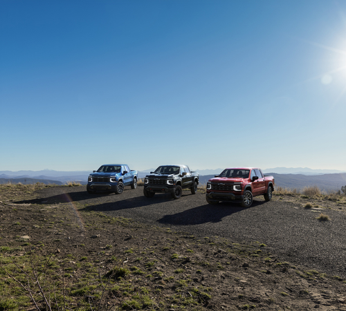 Three pickup trucks in blue, black, and red parked on a scenic hilltop under a clear sky.