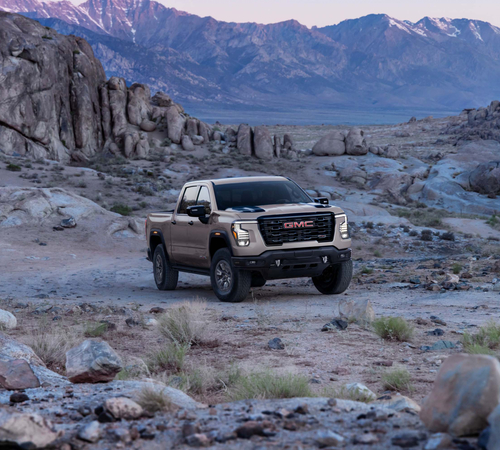 A GMC truck navigates rocky terrain with mountain scenery in the background under a twilight sky.