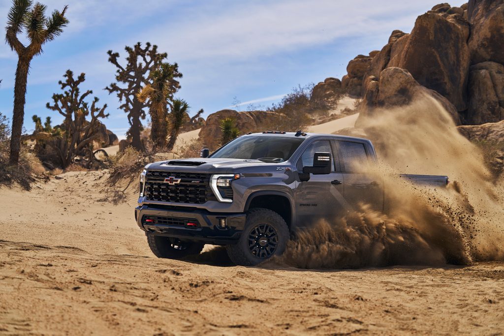 A rugged pickup truck powers through a sandy desert landscape with dust flying, surrounded by Joshua trees.